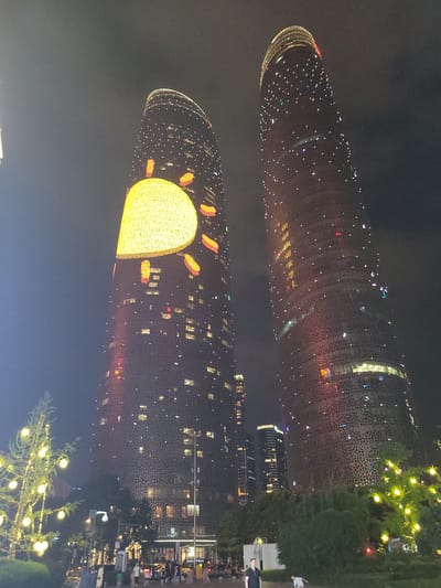 This is a nighttime, ground-level, wide shot of the city of Wuhou District, China, featuring two prominent skyscrapers adorned with lights. The skyscraper on the left has a large, illuminated yellow sun design on its facade, with orange flame-like shapes around it. Both skyscrapers are decorated with numerous small, white, triangular lights, resembling stars. In the foreground, a decorated Christmas tree is visible on the left, also illuminated with yellow lights. The background shows other buildings and streetlights. Several people are present in the scene, walking on a street and sidewalk area, with vehicles visible in the background. The overall atmosphere is festive and urban.