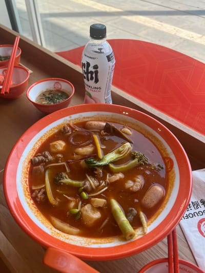 This is a close-up, overhead shot of a bowl of spicy soup with various ingredients, including what appear to be beef, shrimp, broccoli, and tofu, served in an orange-rimmed bowl. There are also two small bowls of dipping sauce or condiments, chopsticks, and a bottle of "Chi Forest" sparkling white peach flavored drink on a wooden table. The scene appears to be indoors, with natural light coming from a window, suggesting a restaurant or eatery. The background is blurred but shows a red surface with a geometric pattern, possibly a table runner or placemat. The overall impression is of a meal being prepared to be eaten. This image was taken at "Malaysian & Chinese Food" restaurant at Prosper-Allee 2 in Bottrop, Germany.