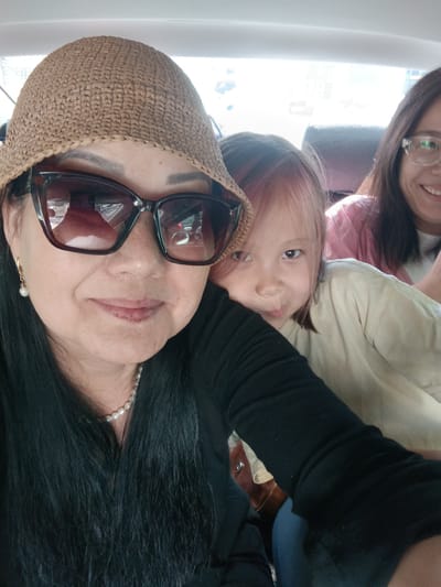 The image shows three individuals inside a vehicle, presumably in the city of Yahşihan, Turkey. In the foreground, a woman wearing a straw hat and large sunglasses smiles at the camera. She has long, dark hair and is wearing a black shirt and a pearl necklace. To her right, a young girl with light-colored hair leans in, making a playful face. Behind them, another woman with dark hair and glasses is partially visible, smiling. The background suggests an interior of a car, with seat upholstery and a window showing a blurry outdoor scene, possibly urban.