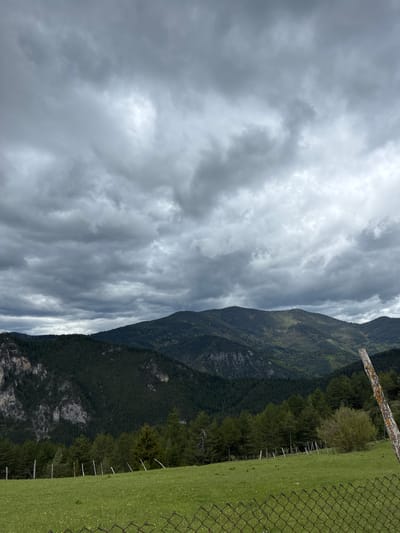 The image shows a mountainous landscape under a cloudy sky. The photograph appears to be taken from a high vantage point, potentially near a meadow with a wire fence visible in the foreground. The mountains are densely forested. The sky is overcast with various cloud formations. The location is Giovren, Bulgaria.