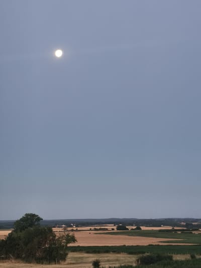 The image shows a wide, panoramic view of a rural landscape at dusk. The sky is a pale, dusky blue, with a bright full moon visible in the upper left quadrant. The moon is round and appears to have a slight halo effect around it. Below the sky, a band of dark green trees runs along the horizon, dotted with a few clusters of buildings that suggest a small village or hamlet. In the foreground and middle ground, there are fields of harvested crops, appearing as a patchwork of golden brown and green. Some scattered trees and bushes are also present in the foreground. The overall impression is one of tranquility and natural beauty in a rural setting. The location is identified as Époisses, France, indicating the geographical context of this serene landscape.