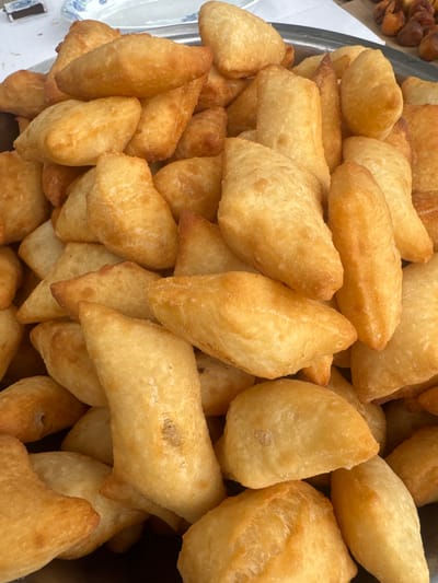 The image displays a close-up, overhead view of a large pile of golden-brown, fried dough pastries. The pastries are irregularly shaped, with some appearing triangular or oval, and others more rectangular. They are stacked and overlapping, filling a metallic bowl. The texture appears slightly puffy and crispy. The background shows hints of white plates and a blurred object, possibly food, in the upper right corner. The scene is indoors, likely a food stall or a home setting, and is not specifically tied to the city of Batken, Kyrgyzstan, though such pastries are common in Central Asian cuisine. There are no people, actions, or interactions depicted.
