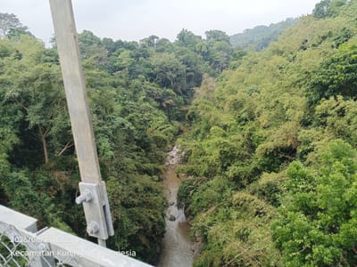 The image shows a view from a bridge overlooking a river valley densely covered with lush green trees and vegetation. A metal support structure of the bridge, with visible bolts and nuts, is in the foreground on the left. A brown river flows through the bottom of the valley, with rocks visible in its course. The trees form a thick canopy covering the slopes of the valley, with some appearing to be bamboo. The sky is overcast, suggesting a cloudy day. The timestamp "2026/06/23 12:23" and the text "Kecamatan Kuningan, Indonesia" are superimposed on the lower left corner of the image, indicating the location and date the photograph was taken. There are no people visible in the image.