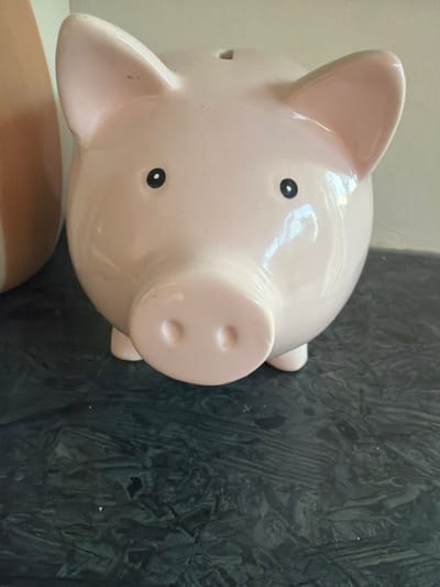 A close-up, eye-level shot shows a pink ceramic piggy bank with black eyes and a snout. The piggy bank is positioned on a dark, textured surface, possibly a countertop. To the left of the piggy bank, a portion of a tall, light-colored object, likely a vase, is visible. The background consists of a plain, off-white wall. The lighting is bright and appears to be artificial. No people are present in the image. The scene does not depict any specific location in Turnhout, Belgium.