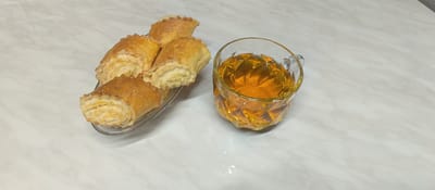 A clear glass cup filled with a golden-amber colored beverage, likely tea, sits on a light-colored marbled surface. The cup has a faceted diamond pattern and a small handle. To the left of the cup is a clear glass serving dish containing four golden-brown pastries. The pastries are crescent-shaped and appear to be made of a flaky dough, rolled and dusted with sugar or a similar sweet topping. The filling is visible in the cut ends of the pastries, appearing as layers of light yellow. The overall scene suggests a moment of relaxation or hospitality, with a sweet treat and a beverage. The setting is indoors, on a clean surface, likely a table. There is no indication of time of day or weather. There is no visible text in the image.