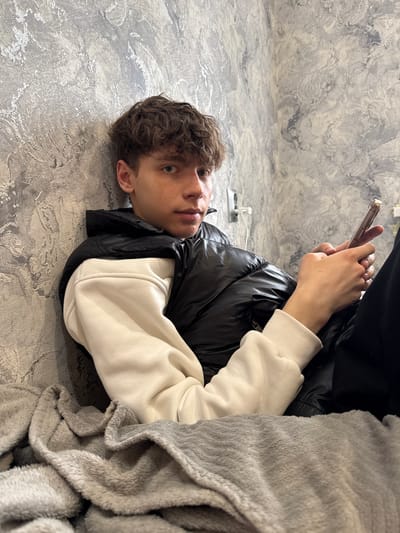 A young man is lying on a textured gray floor, potentially made of concrete or tile. He is wearing a black puffer vest over a light-colored sweatshirt. A gray blanket partially covers his body. He is holding a smartphone in his hands. The background is a gray wall with a mottled, stone-like pattern. An electrical outlet is visible on the floor near the wall. The photo does not contain sufficient information to confirm the specific location as Korobovo, Russia.