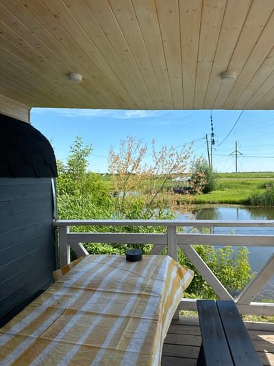 The image shows a covered porch with a wooden ceiling and floor. A table covered with a yellow and white striped tablecloth is in the foreground, with a small dark lantern on it. To the left of the table is a dark wooden structure with a dark, shingled, domed roof. Beyond the porch railing, there is a body of water, green vegetation, and in the background, power lines and poles are visible against a clear blue sky. The scene suggests a rural or semi-rural setting, possibly a dacha or a lakeside retreat. The location is identified as Kommunar, Russia. There are no people visible in the image.