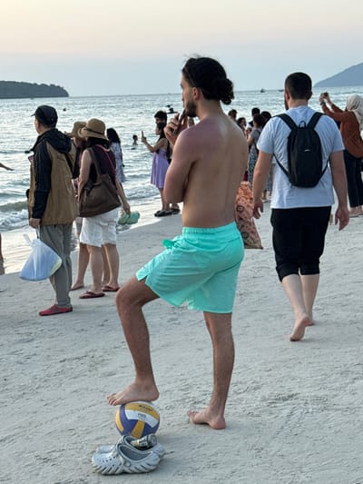 The image depicts a beach scene at Cenang Beach, Malaysia, at what appears to be dusk or early evening. The foreground is dominated by a sandy beach, traversed by numerous individuals.

A shirtless man, with dark curly hair tied back in a bun, is positioned in the lower-left to central frame, facing slightly right, with his back partially visible. He wears mint green swimming shorts. His left foot rests barefoot on top of a yellow, blue, and white volleyball, which in turn rests on a pair of light grey Crocs clogs. A metal beverage can is visible inside one of the clogs. The man's skin on his back shows signs of sunburn. He appears to be holding a dark, elongated object, possibly an electronic cigarette or vape device, to his mouth.

To the left of the central man, a person in a brown jacket and light trousers holds a white plastic bag. Further left, a person in a straw hat and white shorts carries light green flip-flops. Multiple other individuals are scattered along the shoreline and in the shallow water, many appearing to observe the surroundings or take photographs with smartphones. One person, partially obscured, wears a purple dress and holds a phone up. Another person walks away from the viewer in the mid-right, wearing a light blue t-shirt and dark shorts, with a small black backpack. A person in a brown top and head covering is visible in the far right background, also appearing to take a photo.

The background features the calm ocean with gentle waves lapping the shore. A distant island or landmass is visible on the horizon under a pale, overcast or sunset sky. Several small boats are faintly discernible on the water.