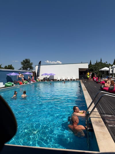 A sunny day at an outdoor swimming pool in Vetren Dol, Bulgaria, shows people swimming and lounging. The pool water is a bright blue. On the left, two women are swimming. Further back, several people are seated on beanbag chairs under purple umbrellas, some with drinks. A man in a yellow vest is walking near the edge of the pool. On the right side of the frame, people are relaxing on large pink inflatable rings. A wooden deck surrounds the pool. In the background, white buildings and trees are visible under a clear blue sky.