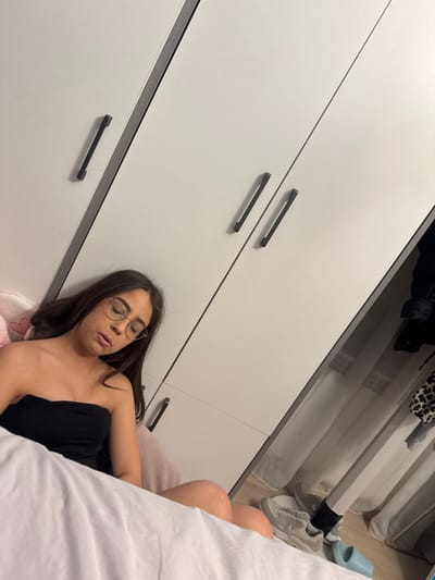 The scene depicts an interior room in the city of Kanchevo, Bulgaria. A female individual with long dark hair, wearing light-framed glasses and a black strapless top, is shown resting or asleep. Her head is supported by a pink cushion or pillow. White bedding partially covers her lower body in the foreground. Behind her, a multi-panel white wardrobe with black horizontal handles occupies the upper portion of the frame. To the right, an open closet section reveals dark hanging garments, including one with a leopard print. On the light-colored floor directly beneath the open closet, a pair of light grey sneakers and a single light blue slide sandal are visible.