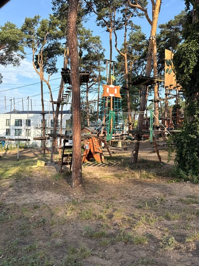 The image shows an outdoor ropes course in the city of Sława, Poland. The course is set among tall pine trees and features various obstacles, including wooden platforms, rope bridges, and nets. A life preserver hangs from a tall structure in the center of the image, which appears to be part of the course. In the background, a modern white building with solar panels on its roof is visible. The ground is covered in sand and sparse vegetation. The sky is clear and blue. There are no people visible in the image.