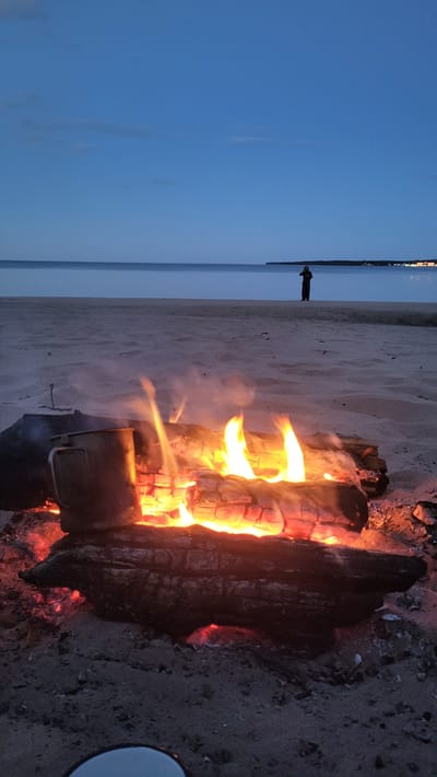 A campfire burns on a sandy beach at dusk, near the village of Zaostrov'e, Russia. The fire, made of several logs, emits bright orange flames and smoke. A metal mug sits to the left of the fire, on a log. In the background, a calm sea stretches to the horizon under a deep blue sky with a few wispy clouds. A lone figure, silhouetted against the sky, stands on the beach in the distance, facing the water. The scene suggests a tranquil evening by the coast.