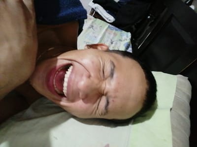 The scene depicts a close-up, tilted view of an indoor setting, located in Soacha ciudad, Colombia. The primary subject is an adult male, shown from approximately the mid-chest upward. He has short, dark hair. His eyes are closed, and his facial muscles are contracted, creating deep creases around his eyes and cheeks. His mouth is open wide, revealing both upper and lower teeth. A gold-colored chain is visible around his neck. To his left shoulder, a folded piece of dark blue fabric is present. He is positioned on a light green pillow. In the background, to the upper right, crumpled white fabric with cartoon characters is visible, alongside other dark clothing items. A dark, possibly wooden or laminate, furniture piece is partially discernible in the far upper right background. The lighting is ambient.