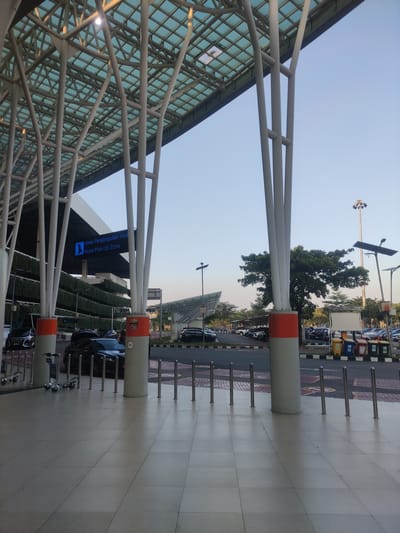 The image displays the exterior of a modern airport terminal or similar transit hub, specifically a "Hotel Pick-Up Zone," located in Rawa Burung, Indonesia.

The dominant feature is a large, contemporary canopy structure covering a pedestrian area. This canopy comprises a grid of translucent or transparent panels, likely glass, supported by numerous tall, branching white structural columns. Several of these columns have prominent orange bands at their lower sections. Sunlight filters through the canopy and casts shadows on the ground.

To the left, underneath the canopy, is a multi-story building featuring a facade partially covered in tiered horizontal planters overflowing with lush green foliage, creating a "green wall" effect. A prominent blue digital sign hangs from the building, displaying white text that reads "Area Penjemputan Hotel / Hotel Pick-Up Zone" alongside an icon of a person with luggage.

In the mid-ground, beyond the central columns, a paved roadway separates the pedestrian area from a large parking lot. Several black cars, including sedans and SUVs, are visible on the roadway and parked near the building. In the distance, within the parking lot, numerous other vehicles of various colors are parked under partial cover structures. The parking lot also features tall streetlights, one with a large array of floodlights and others with solar panels, along with scattered trees. Multiple segregated waste bins (yellow, blue, green) are visible near a blank white sign in the parking lot.

The immediate foreground consists of light-colored, square-tiled pavement. Rows of polished silver bollards (metal posts) are installed on the pavement, serving as a barrier between the pedestrian zone and the vehicle area. Two silver luggage trolleys are visible on the left side of the pedestrian path. The sky above is clear and light blue, indicating daytime conditions. No people are discernibly visible within the frame.