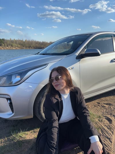 A woman in a white shirt, black blazer, and sunglasses is lying on the hood of a silver car. The car is parked on a dirt surface near a body of water. The sky is blue with scattered clouds. The location is near a body of water in Zaosinovo, Russia.