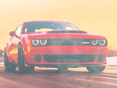 The image is a low-angle, close-up, head-on shot of a red Dodge Challenger SRT. The car is positioned in the foreground, with its front grille and headlights prominently displayed. The background is out of focus and appears to be a sunset or sunrise with a warm, orange hue. There are subtle, light-colored cracks visible across the surface of the image, suggesting it may be a photograph of a print. The car's wheels and part of its side are visible. The car is stationary, and there are no people or other objects clearly depicted in the scene. The lighting suggests a natural light source. There is no discernible connection to Paczkowo, Poland, in the provided image.