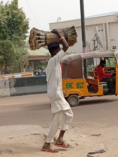 The image displays an outdoor street scene in Darmanawa, Nigeria, under bright daylight conditions. In the foreground, a dark-complexioned male walks from left to right on a dirt shoulder beside a paved road. He wears a light-colored traditional tunic and trousers, a dark blue cap, and pink slide sandals. He balances a large, cylindrical bundle of natural fiber brooms, secured with black straps, on his head, with his left hand slightly raised near the load. Behind him, on the paved road, a yellow three-wheeled motorized rickshaw, identified by "KMC 1027" and "SPORT" markings on its side, is visible. Inside the rickshaw, a second dark-complexioned male, dressed in a red long-sleeved shirt and dark trousers, is seated at the controls. The background includes green trees and shrubs, a grey concrete road barrier, and a light-colored building equipped with multiple satellite dishes. A dark utility pole stands vertically in the midground, and a white container bearing the text "ENEOS" is partially visible to the left of the scene.
