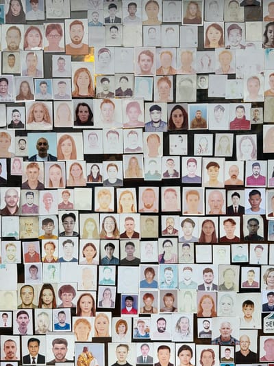 A wall in Ghandruk, Nepal, displays a dense, irregular collage of numerous individual portrait photographs and sketches. These portraits, predominantly headshots and bust compositions, depict a diverse array of individuals varying in age, gender, and ethnicity. Many of the images exhibit signs of wear, fading, water damage, or partial obscuration; some are photographic prints while others appear to be hand-drawn or sketched. The photos are haphazardly affixed, frequently overlapping, onto a dark background surface. This display likely serves as a record or memorial for visitors or trekkers in a lodge or teahouse.