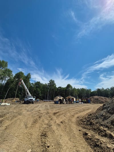 A construction site is depicted under a bright blue sky with wispy clouds. The foreground is dominated by a dirt track with tire marks. In the mid-ground, a white crane truck is parked on the muddy ground. To the right of the crane, several portable structures or sheds are visible, some with open doorways. A pile of reddish-brown earth is located to the right of these structures. In the background, a dense forest of green trees lines the horizon. Several individuals in yellow vests are present on the site, engaged in work or observation. The scene appears to be a construction project in progress in a rural or undeveloped area, possibly near Taezhnoe, Russia, given the mention of the city.