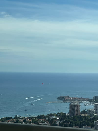 The image is an aerial view of a coastal city, likely Vrela, Montenegro, on a clear day with scattered clouds. The deep blue sea occupies the majority of the frame, with the horizon clearly defined. Several boats are visible on the water, some leaving white wakes, indicating movement. In the distance, a red parachute is visible, suggesting recreational activity. The coastline is lined with buildings, including residential areas and a prominent harbor filled with yachts and smaller vessels. A historic walled town with red-tiled roofs and a church spire is situated on a peninsula jutting into the sea. In the foreground, modern buildings, including a high-rise under construction, are visible alongside lush green trees. A dark railing runs along the very bottom edge of the frame, suggesting the photo was taken from a high vantage point.
