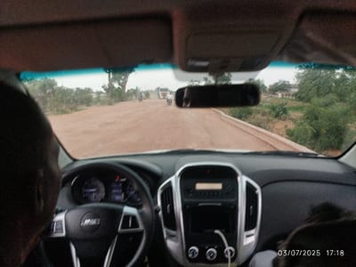 The view from inside a car shows a dirt road ahead. The car's dashboard is visible with the steering wheel to the left. A rearview mirror hangs above the dashboard, reflecting the road behind. Trees and vegetation line both sides of the road. In the distance, a motorcycle and a white truck can be seen. The date "03/07/2025" and time "17:18" appear in the lower right corner.