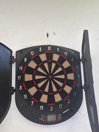 The image shows a black electronic dartboard with its protective cabinet partially opened. The dartboard itself is mounted on a white, textured wall. The dartboard features the standard numbered segments from 1 to 20, with red and black alternating sections, along with the bullseye. The electronic component of the board, located on the right side, has a screen and several buttons for game control. The image was not taken in the city of Slashten, Bulgaria.