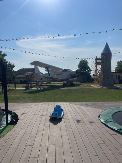 In the city of Zîrnești, Moldova, on a sunny day with a clear blue sky and scattered clouds, an outdoor area features a unique display. A biplane, painted white with blue accents, is parked on a grassy field. Nearby, a tall, grey, conical structure resembling a rocket stands next to a wooden ladder. A string of colorful triangular flags is strung across the sky above the biplane and rocket. In the foreground, a wooden deck is visible, with a blue children's ride-on toy and the edge of a green-padded trampoline. Three young boys are walking on the grass near the biplane. The environment suggests a recreational space, possibly a park or a children's attraction.