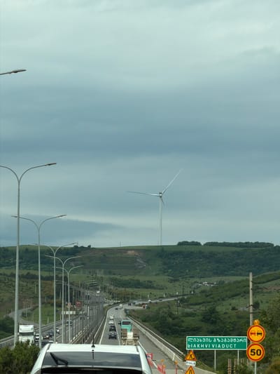 The image shows a highway in Tedotsminda, Georgia. The camera is positioned on the road, looking towards a wind turbine. The sky is overcast. Alongside the road is a grassy hill. Traffic is visible on the highway. Road signs are present. Streetlights line the road. A vehicle is partially visible in the foreground, presumably the source of the image.