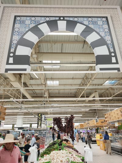The image depicts the interior of a large supermarket. The upper portion shows a decorative archway with intricate tile patterns in blue and white, and gray and white geometric shapes. Below the archway, the supermarket aisles are visible, illuminated by fluorescent lighting. People are seen shopping in the aisles, with displays of fresh produce like garlic, onions, eggplants, and gourds prominently featured in the foreground. Various refrigerated units and shelving units stocked with products are also visible. A sign in Arabic and French indicates a price of 7.90. The overall environment appears to be a well-stocked, modern retail space.