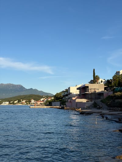 The image shows a coastal scene in Krašići, Montenegro. The view is divided between a clear blue sky on the left and the Adriatic Sea on the right. A line of buildings and mountains runs along the horizon, separating the sky and sea. Closer to the top, construction is visible near the water.