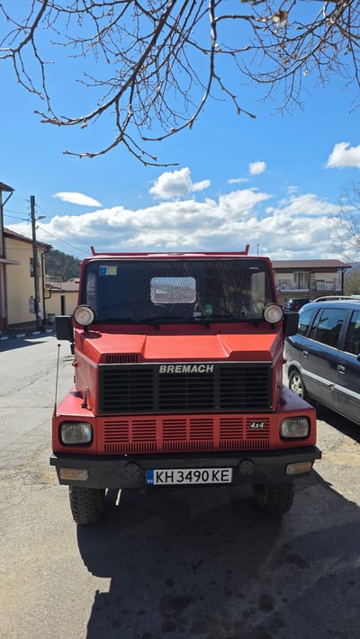 A red Bremach 4x4 vehicle with the license plate KH 3490 KE is parked on an asphalt surface. The front of the vehicle is visible, showing the BREMACH logo. A dark-colored car is partially visible behind the Bremach. Buildings are visible in the background. The sky is blue with scattered clouds. Branches of a tree are in the left of the frame. The image was taken in Slatino, Bulgaria.
