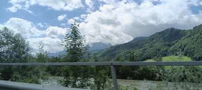 The image shows a scenic view of a river valley with lush green mountains in the background. A silver railing is visible in the foreground, suggesting the photo was taken from a moving vehicle. The sky is partly cloudy with dramatic formations of white and grey clouds. A river with slightly turbulent water flows through the valley, bordered by dense green foliage and trees. In the middle ground, a tall, slender tree stands out against the backdrop of the mountains. The mountains are covered in thick forests, with some rocky peaks visible in the distance. The overall impression is one of natural beauty and tranquility. This scene is likely located near Hallein, Austria, given the characteristic Alpine landscape.