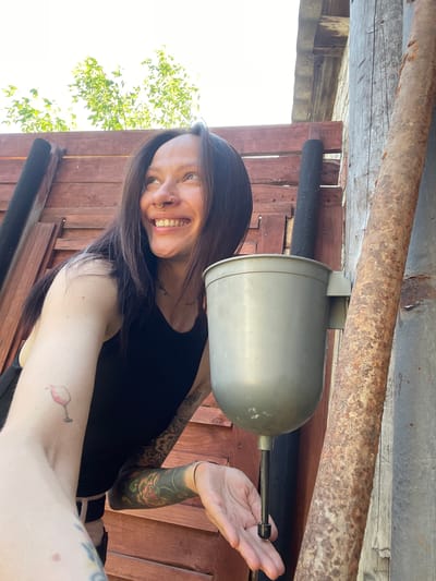 A photograph depicts a woman standing beneath a weathered, open-air structure, possibly a shed or shelter. The woman is fair-skinned with dark hair and visible tattoos on her arms. She is smiling, and her hand is extended toward a gray, cylinder-shaped object hanging from the structure. The structure's ceiling is constructed of wood and metal piping that displays signs of rust. Wooden planks form the wall behind her, and a glimpse of sky and foliage is visible in the lower left of the frame. The location is not evident from the image, and therefore the information about Chemoshur, Russia, cannot be confirmed from the visual data.