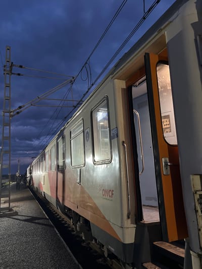 The image displays a Moroccan National Railways (ONCF) passenger train stopped at a station platform in Sidi Slimane, Morocco, during twilight or night conditions. The train, positioned on tracks adjacent to a dark platform, features a white body with red and orange livery; the "ONCF" logo is visible on its side. Interior lights illuminate the train's windows and an open passenger door near the foreground, revealing interior steps and a corridor. A number "2" is affixed near a window on the visible side of the train. Overhead, catenary lines are suspended from a concrete support structure against a dark blue, cloudy sky. Several indistinct human figures are visible on the platform further along the train.