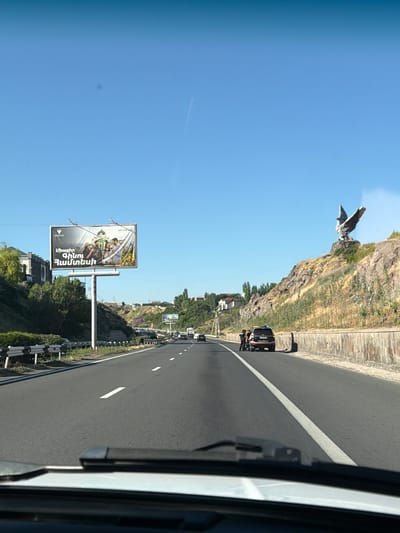This is a ground-level, eye-level, medium shot taken from inside a vehicle looking forward on a highway on a clear, sunny day. The highway has at least two lanes in each direction, separated by a white line. A highway guardrail is visible on the left side of the road.

On the left side of the highway, there is a billboard with an advertisement featuring three people and a building. Further up the embankment, some trees and buildings are visible.

On the right side of the highway, a large eagle statue is perched on a rocky outcropping overlooking the road. In front of the statue, a dark-colored SUV is parked on the shoulder, and three individuals, two in dark uniforms and one in lighter clothing, are standing near the back of the vehicle.

The sky is a clear blue with a faint contrail visible. The overall scene depicts a highway leading through a semi-urban or suburban area with both natural and man-made landmarks. The location is Getamej, Armenia.