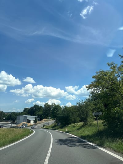 The image depicts a sunny day with a bright blue sky dotted with white cumulus clouds. A paved road curves to the right, bordered by grassy verges and trees. On the left side of the road, there is a building complex with a grey, modern structure and some piles of wood chips. Two white cars are visible on the road, one approaching the curve and another further ahead. The right side of the road is lined with lush green trees and grass. A signpost with text in a foreign language is visible on the right side, near the trees. This image was captured in Litomyšl, Czechia, likely on a rural road leading out of the city.