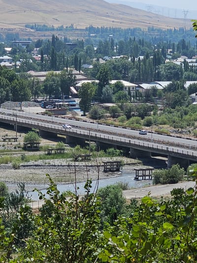 The image depicts a wide, elevated view of a city in Шералы, Kyrgyzstan. A long, multi-lane bridge spans a dry riverbed filled with rocks and a shallow stream. The bridge has a white railing and yellow accents along its edges. Several vehicles, including a white car, are visible on the bridge. Below the bridge, there are concrete structures and benches. The surrounding landscape features a dense collection of trees, houses with white roofs, and some industrial buildings. In the background, a dry, hilly terrain rises, dotted with power lines and transmission towers. The overall scene suggests a sunny day with clear visibility.