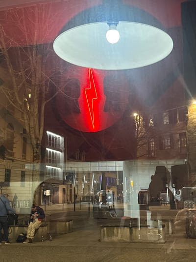 A white lamp with a lightbulb shines in front of a red circle containing a red lightning bolt. Behind this is a reflection in a window. Buildings, bare tree branches, a street, and people are reflected. The reflection shows a store interior with tables and chairs. People are on the sidewalk outside the store.