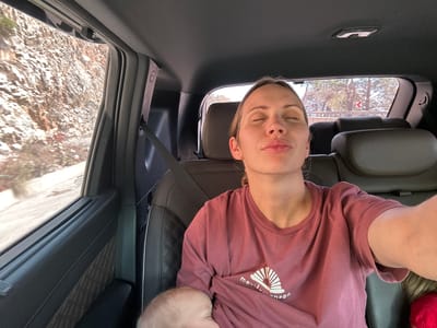 A woman is photographed from inside a vehicle, possibly a car, while nursing a baby. The woman is wearing a burgundy t-shirt and has her eyes closed. She is wearing a seatbelt, suggesting she is a passenger. Through the car windows, a landscape with what appears to be mountainous terrain is visible. The location is suggested to be in the city of Ula, Turkey.