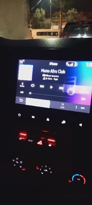An in-car entertainment system displays "Mano Afro Club" by "Album Inconnu" and DJ Velo - Topic. The system's display indicates a temperature of 19°C. A car air conditioning system is visible, and other dashboard buttons with red accents are illuminated. A glimpse of the outside world, showing trees and buildings, is visible through the car's window.