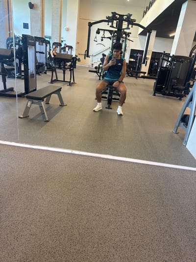 A person sits on gym equipment, seemingly taking a selfie in a mirror. The gym has various weight machines and benches, with beige speckled flooring. The reflection includes a view of the gym's layout, showing additional exercise stations and equipment. The mirror's edge is prominent, separating the reflected gym interior from a solid beige surface.