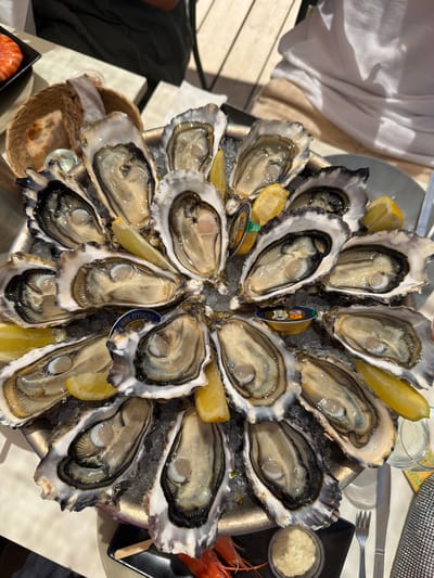 An overhead shot shows a large platter of oysters on ice, arranged in a circular pattern. There are approximately 20 oysters, each served open with lemon wedges. Two small tubs of sauce are visible on the platter, as well as a small glass of what appears to be mignonette. A bread basket containing sliced bread is positioned to the left of the oyster platter. In the background, parts of people can be seen wearing casual clothing. The setting appears to be outdoors, possibly a restaurant terrace, with wooden decking visible. A small black plate with two red shrimp and a small white ramekin of sauce is in the lower left corner of the image. In the upper left corner, there is a small black plate with two cooked shrimp. The location is a restaurant in Lège-Cap-Ferret, France.