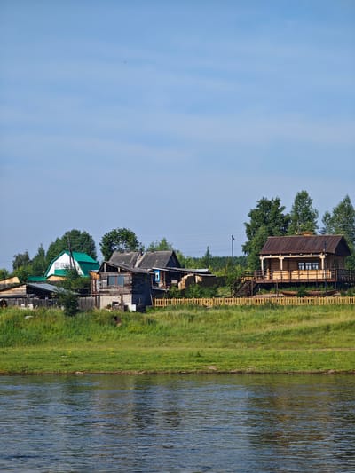 The image depicts a rural village scene in Galanino, Russia, on a clear day. In the foreground, a wide, calm river with a dark, reflective surface dominates the lower half of the frame. The riverbank is lined with lush green grass, which slopes upwards towards a cluster of rustic wooden houses and structures.
The village itself is situated on the elevated bank. Several wooden houses are visible, varying in architectural style and condition. One prominent structure on the right is a well-maintained log cabin with a wooden deck and stairs leading up to it. It has a dark-colored roof and several windows. To the left of the log cabin, there are older, more weathered wooden houses with corrugated roofs, some appearing to be in disrepair. One of these has a distinctive blue-framed window. Various outbuildings, fences, and stacks of lumber are scattered among the houses. A light-colored wooden fence runs along the crest of the bank, separating the village from the river.
The background is filled with dense green trees, forming a natural backdrop to the settlement. The sky is a clear, bright blue with a few wispy clouds and a distant, tiny dark speck that could be a bird. The overall atmosphere is one of peaceful rural life, with the river and the surrounding nature playing a significant role in the setting.