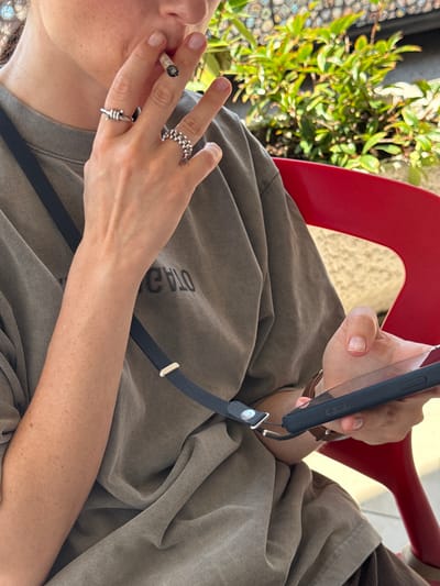 The media file shows a close-up of a person sitting in a red chair, holding a cigarette and a smartphone with a lanyard. The person is wearing a loose-fitting brown shirt. The foreground focuses on their hands, one of which is holding the cigarette, and the other is holding the phone. The person is also wearing rings on their fingers. In the background, there are green plants and a textured surface that appears to be a wall or ground. The lighting suggests it is daytime, possibly outdoors, and the overall mood appears relaxed. There is text visible on the shirt, partially obscured, which appears to read "OTA" and possibly more letters. The setting is Liješnje, Montenegro.