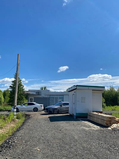 The image depicts an outdoor scene under a clear blue sky with scattered white clouds. In the foreground, a gravel driveway leads towards several structures. To the left, a white sedan is parked alongside a building. To the right of the white sedan, a grey sedan is parked in front of a small, white, rectangular structure with a green trim on its roof. A stack of lumber is neatly arranged to the right of this structure. Behind the sedans, a long, single-story building with a light-colored facade and a dark grey lower section is visible. This building has several windows covered with security bars. A wooden utility pole stands to the left of the buildings, with electrical wires extending from it. Lush green trees and vegetation surround the structures, suggesting a rural or semi-rural setting. The presence of lumber indicates potential construction or renovation activities. The overall scene appears to be a commercial or storage area.