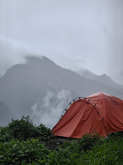 An orange tent is pitched on a rocky slope covered with green vegetation. The tent is partially visible, with its entrance and side facing the viewer. The background features a mountain range shrouded in mist and clouds, indicating a remote and possibly cold environment. The sky is overcast and gray. The image was taken in Dhundi, India.