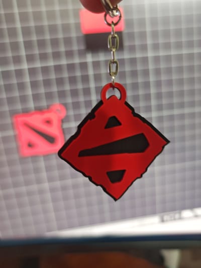 A red and black keychain featuring the Dota 2 logo hangs from a silver chain. The logo is a stylized 'D' within a diamond shape. The keychain is partially out of focus, with a grid pattern visible in the background, possibly a cutting mat. Another blurred, red Dota 2 logo is visible in the background. A person's finger is visible at the top left corner holding the chain. The scene appears to be indoors, with a desk or table in the foreground.