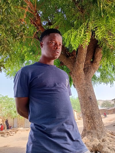 An adult male is standing under a large tree in Nata-Ala, Nigeria. He is wearing a dark blue t-shirt. The tree has dense green foliage. In the background, there is a building and a road. A person on a bicycle is visible in the background.