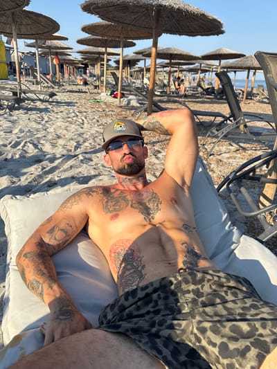 The media file shows a man lying on a cushioned lounger on a sandy beach. He is shirtless and has extensive tattoos covering his chest, arms, and abdomen. He is wearing leopard-print shorts and a baseball cap. Sunglasses are perched on his nose, and he is looking towards the camera. The sun is shining brightly, casting shadows on the sand, indicating it is daytime. In the background, rows of thatched umbrellas and beach chairs suggest a resort or public beach area. The sea and the sky are visible in the distance. The overall impression is one of relaxation and leisure in a sunny, coastal environment. The location context provided is Moudros Municipal Unit, Greece. There is text visible on the man's cap: a circular logo with an image and some writing that is not clearly legible.