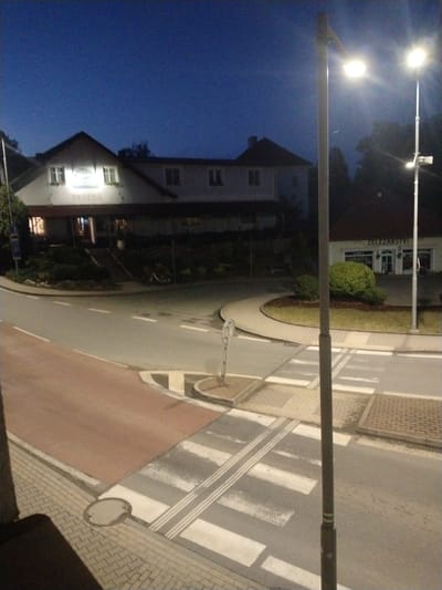 The image depicts a street scene at dusk in Častolovice, Czechia. A two-story building with a pitched roof and arched entrance is visible on the left, with signage reading "DESEDA". Across the street, a smaller building with a flat roof has signage identifying it as "ZELEZARSTVI" (Ironmongery). The street features a pedestrian crossing with tactile paving. Streetlights illuminate the scene. In the background, trees and the night sky are visible. No people or vehicles are present in the immediate scene.